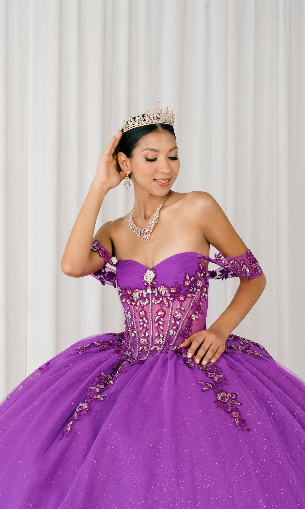 Woman in a Quinceanera Dress 8405JQ By Cinderella Couture with floral applique and a tiara poses in front of white curtains, holding the tiara with one hand and resting the other on her dress.